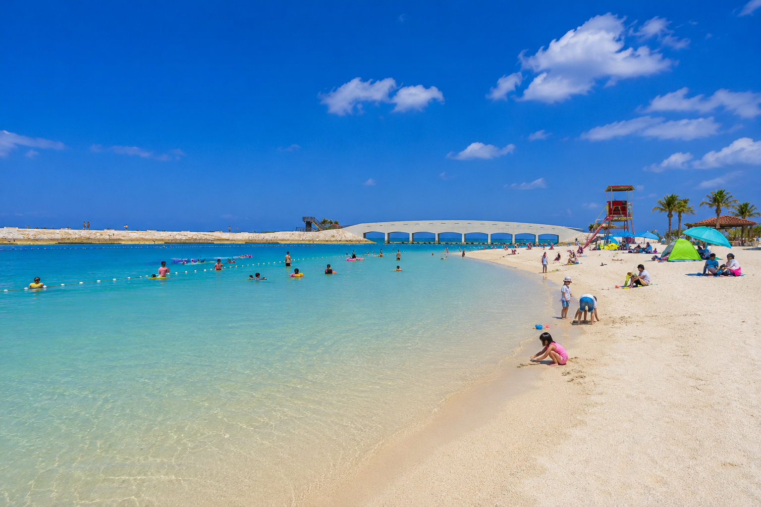 美々ビーチの白い砂浜と透明なエメラルドグリーンの海。青空の下、海水浴を楽しむ人々やカラフルなテントが並び、奥にはアーチ状の橋とヤシの木が見える沖縄のビーチ風景。