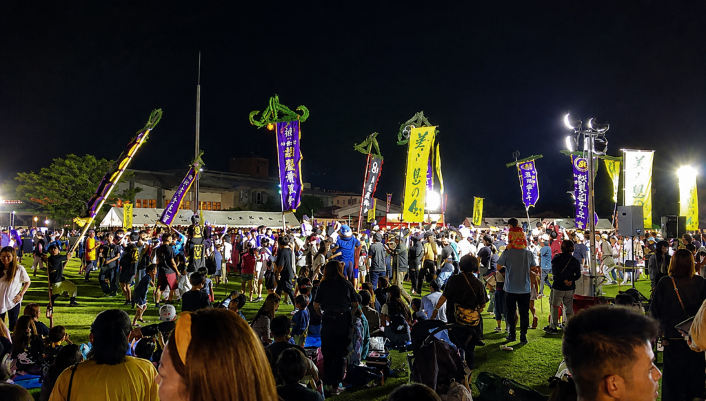 沖縄のエイサー祭りで、夜の会場に色鮮やかな旗やのぼり青年会が立ち並び、多くの人々が芝生の広場に集まっている様子。